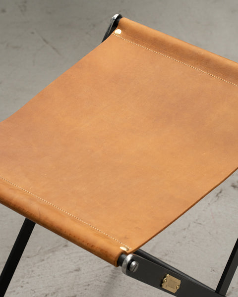 Close-up of a brown leather stool with black metal legs on a concrete floor.